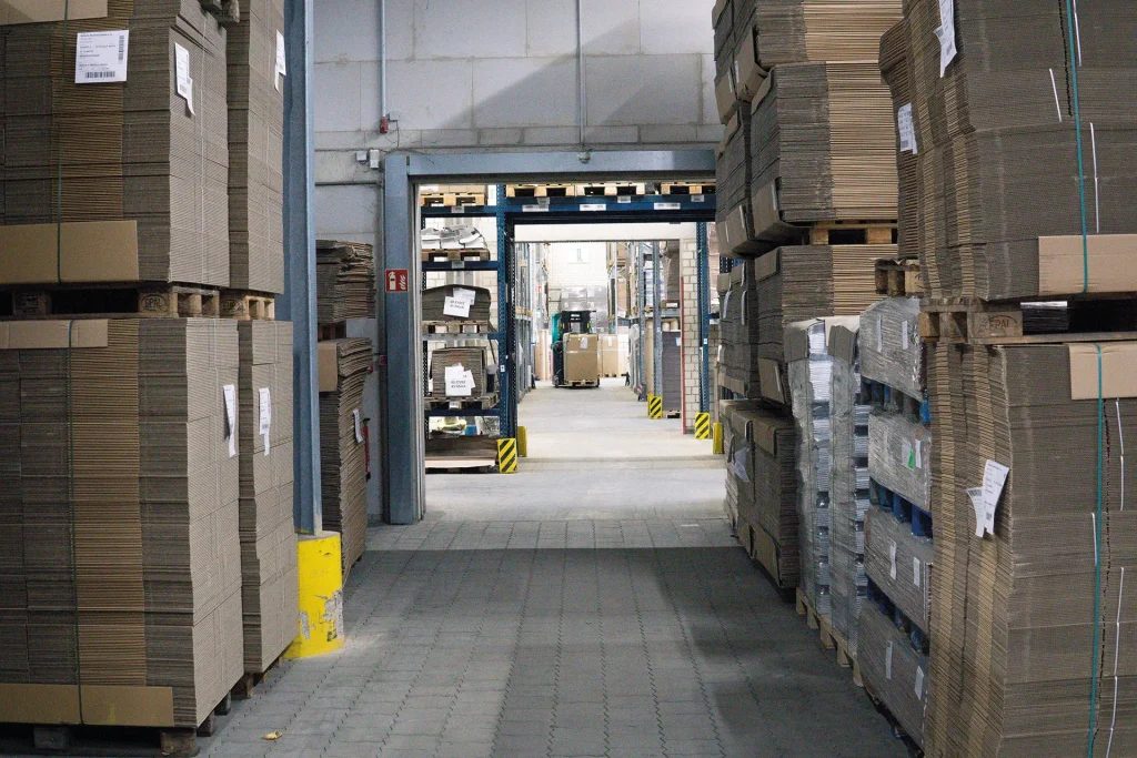 Das Bild zeigt eine Lagerhalle mit gestapelten Kartons. Ein Gabelstapler transportiert Waren im Hintergrund.