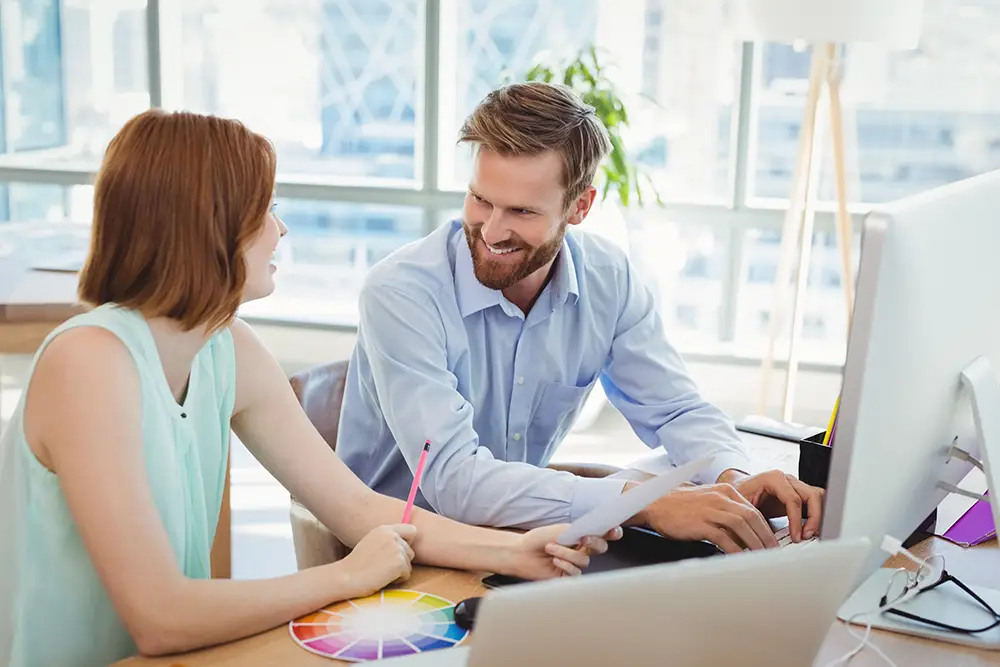 Hummel Papper Beratung: Two colleagues working on a computer with color wheel and documents, smiling at each other, in the office.